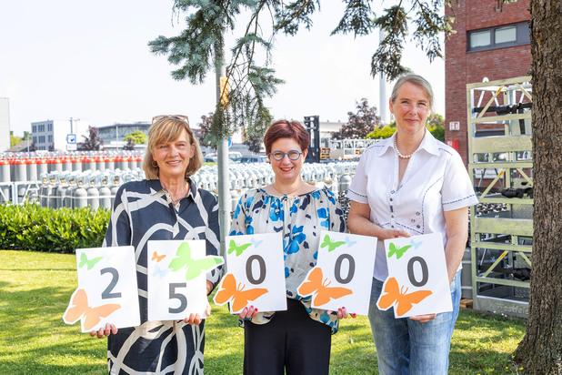 Drei Frauen halten gemeinsam farbige Schilder mit der Zahl 25000 in einem grünen Garten vor einer Gasflaschenlagerung.