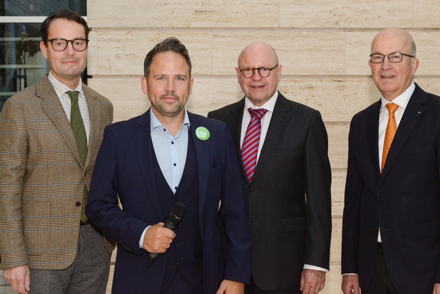 Vier Männer in formeller Kleidung stehen gemeinsam vor einer hellen, strukturierten Wand.