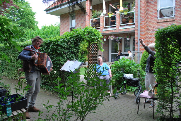 Mann mit Akkordeon spielt im Garten, während Zuhörerinnen entspannt auf Stühlen sitzen und applaudieren.