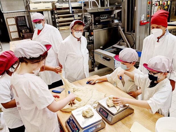 Gruppen von Kindern und Erwachsenen arbeiten in einer Bäckerei, um Teig abzuwiegen und zu bearbeiten.