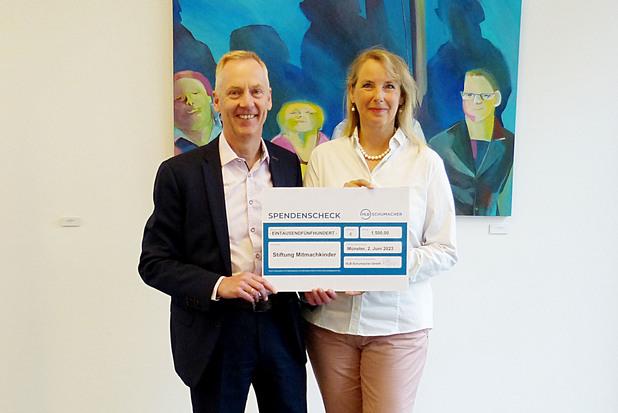 Zwei Personen halten einen Spendenscheck vor bunten Kunstwerken in einem modernen Innenraum.