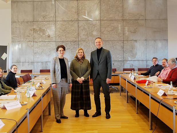 Drei Personen stehen lächelnd vor einer Wand mit reliefartiger Gestaltung in einem Konferenzraum, während Teilnehmer sitzen.
