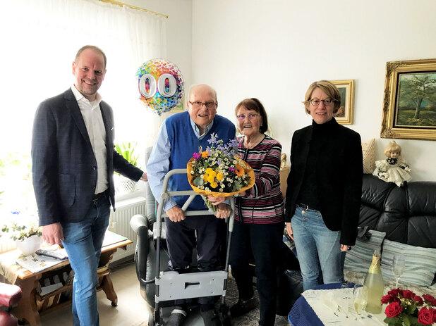 Vier Personen feiern in einem Wohnzimmer einen 100. Geburtstag, der Jubilar hält einen Blumenstrauß und steht an einem Rollator.