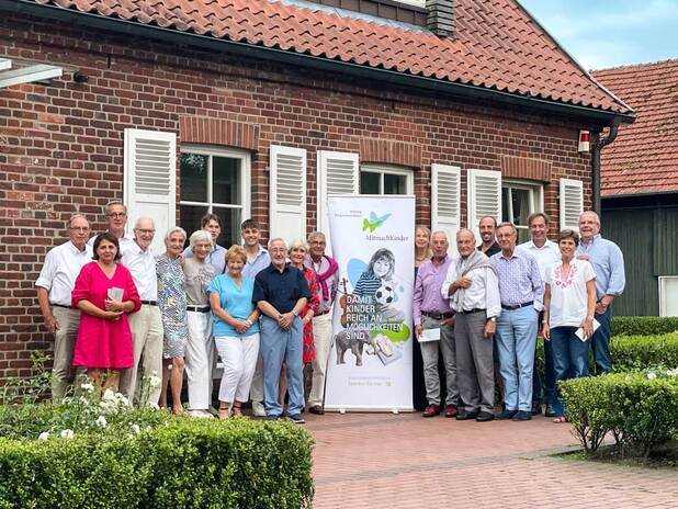 Gruppe von 20 Personen lächelt vor einem Gebäude, neben einem Banner für die Organisation "MitmachKinder".