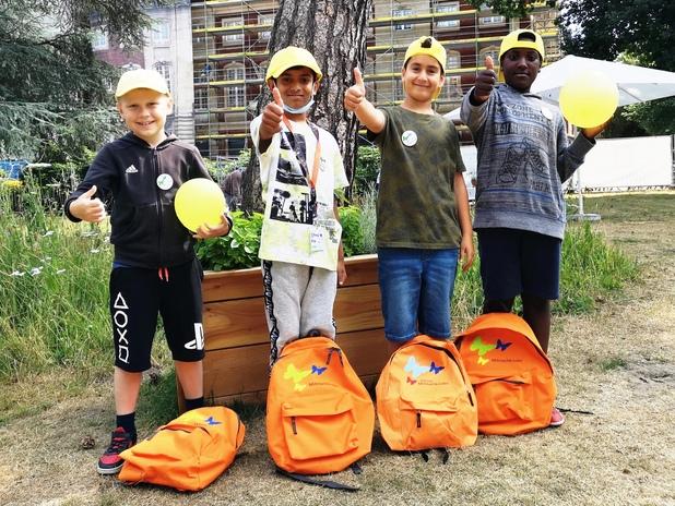 Vier Kinder mit gelben Caps und T-Shirts stehen vor einer Holzbank, zwei halten Luftballons, andere zeigen Daumen hoch.