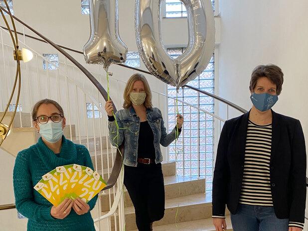 Drei Personen, maskiert, stehen auf einer Treppe und halten silberne Ballons mit der Zahl 10 und eine bunte Plakatkarte.