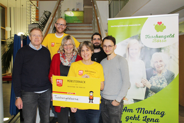 Gruppe von sechs Personen hält ein Schild mit der Aufschrift „Taschendbörse“ vor einem Banner der Taschengeldbörse.