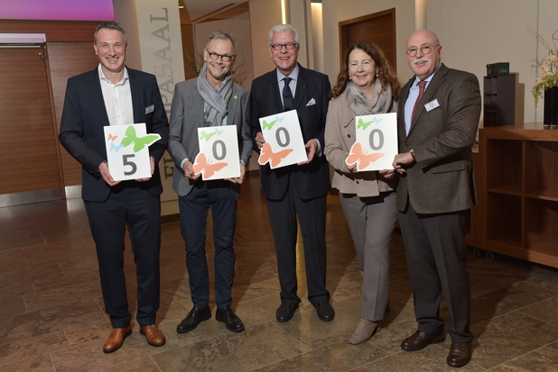 Fünf Personen stehen nebeneinander und halten große Karten mit den Zahlen 5, 0, 0, 0, dekoriert mit Schmetterlingen.