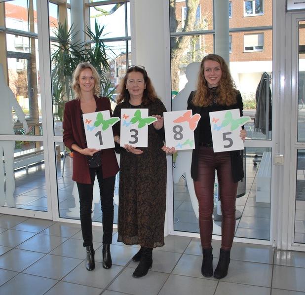 Drei Frauen stehen vor Glasfenstern und halten Plakate mit den Nummern 1, 3, 8 und 5, umgeben von Pflanzen.