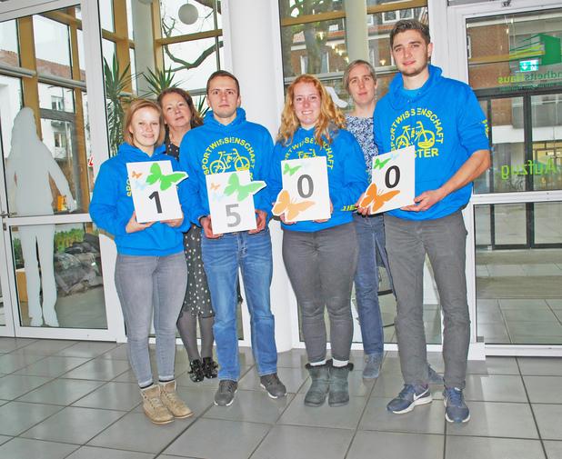 Gruppe von fünf Personen in blauen Sweatshirts hält Zahlen und tierische Motive vor einer Glaswand im Hintergrund.