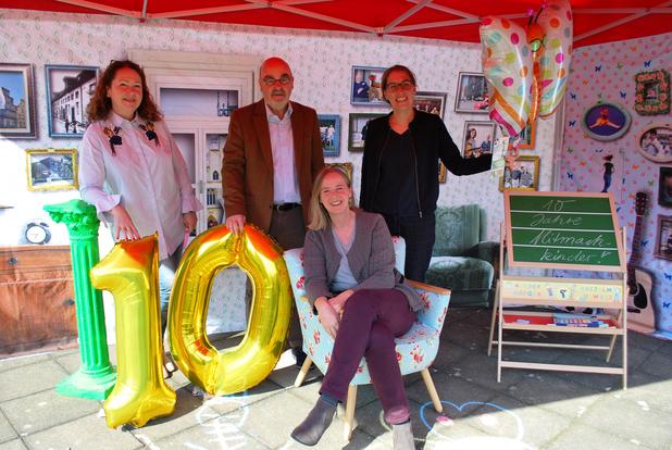 Vier Personen stehen und sitzen unter einem Zelt, neben goldenem Zahlenschild „10“ und einer Tafel mit „10 Jahre Mitmachkinder“.