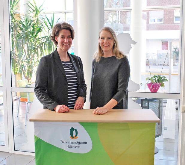 Zwei Frauen stehen an einem Empfangstisch mit einer Tischdecke der Freiwilligen-Agentur Münster, umgeben von Pflanzen.