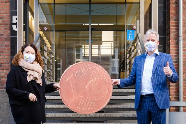 Zwei Personen halten eine große 1-Cent-Münze vor einem Gebäude mit verglaster Eingangstür.
