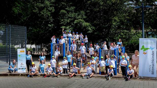 Gruppierung von Kindern und Betreuern auf einem Spielplatz, umgeben von Bäumen, mit einer Werbetafel im Vordergrund.