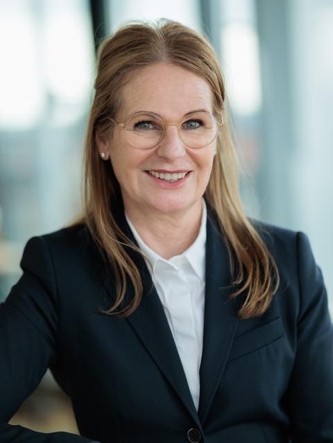 Porträt einer lächelnden Frau mit Brille und blonder, mittellanger Haarpracht in einem schwarzen Blazer.